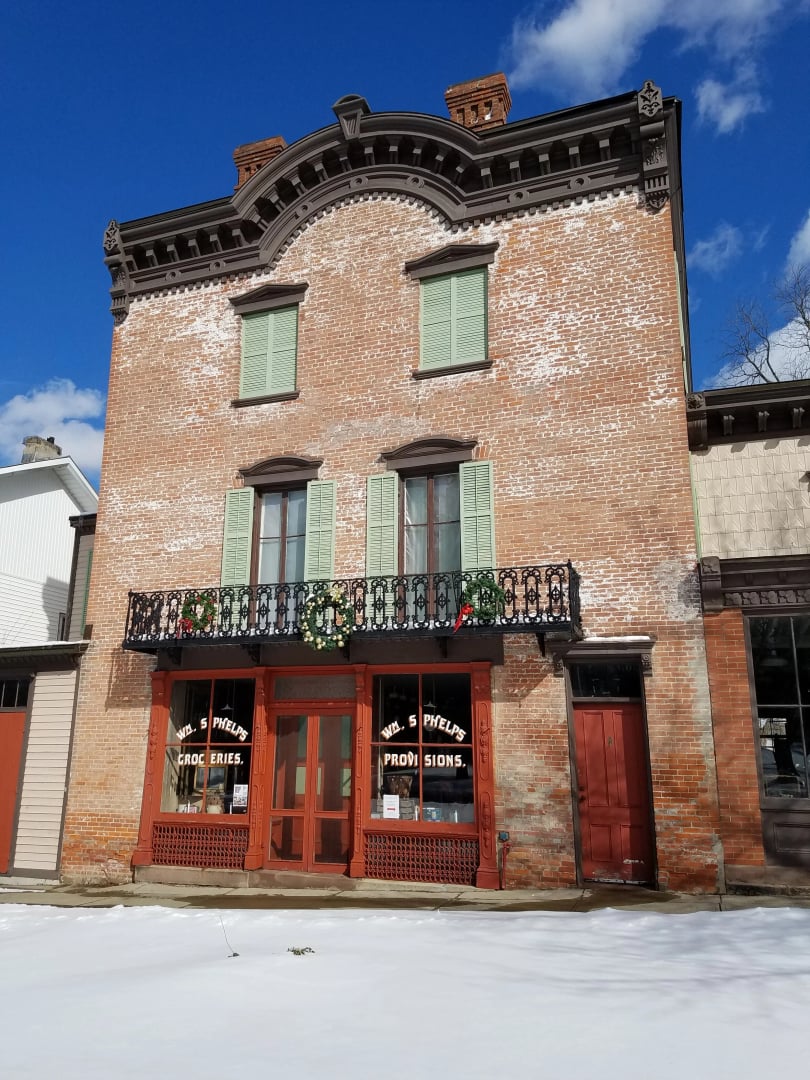 The three-story brick Wm. Phelps General Store on Market Street in Palmyra, New York, with green window shutters and a decorative wrought-iron balcony