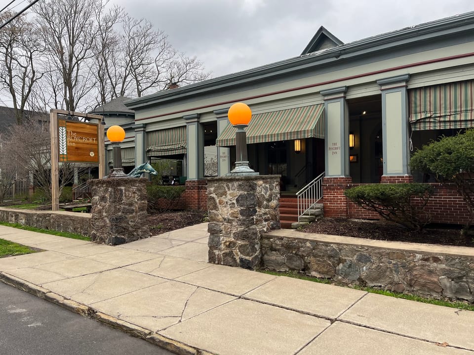 Daytime street-level view of the Roycroft Inn's stone-and-shingle entrance wing with striped awnings and globe lamp posts, East Aurora, New York.