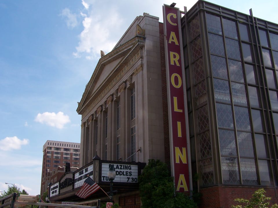 The Carolina Theatre on South Greene Street in Greensboro, North Carolina, seen from a low angle at one end of the front, with its vertical CAROLINA sign, lattice-windowed tower, pedimented stone facade and marquee, in July 2005.