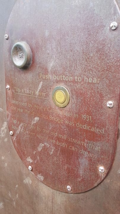 Close up of a sign describing permanent outdoor exhibit of a metal river at Cathedral Park, under the St. John's Bridge in Portland Oregon, installed with music box tune of Hoagy Carmichael's "Up A Lazy River", the year the bridge was built. According to the plaque, it was 1931.