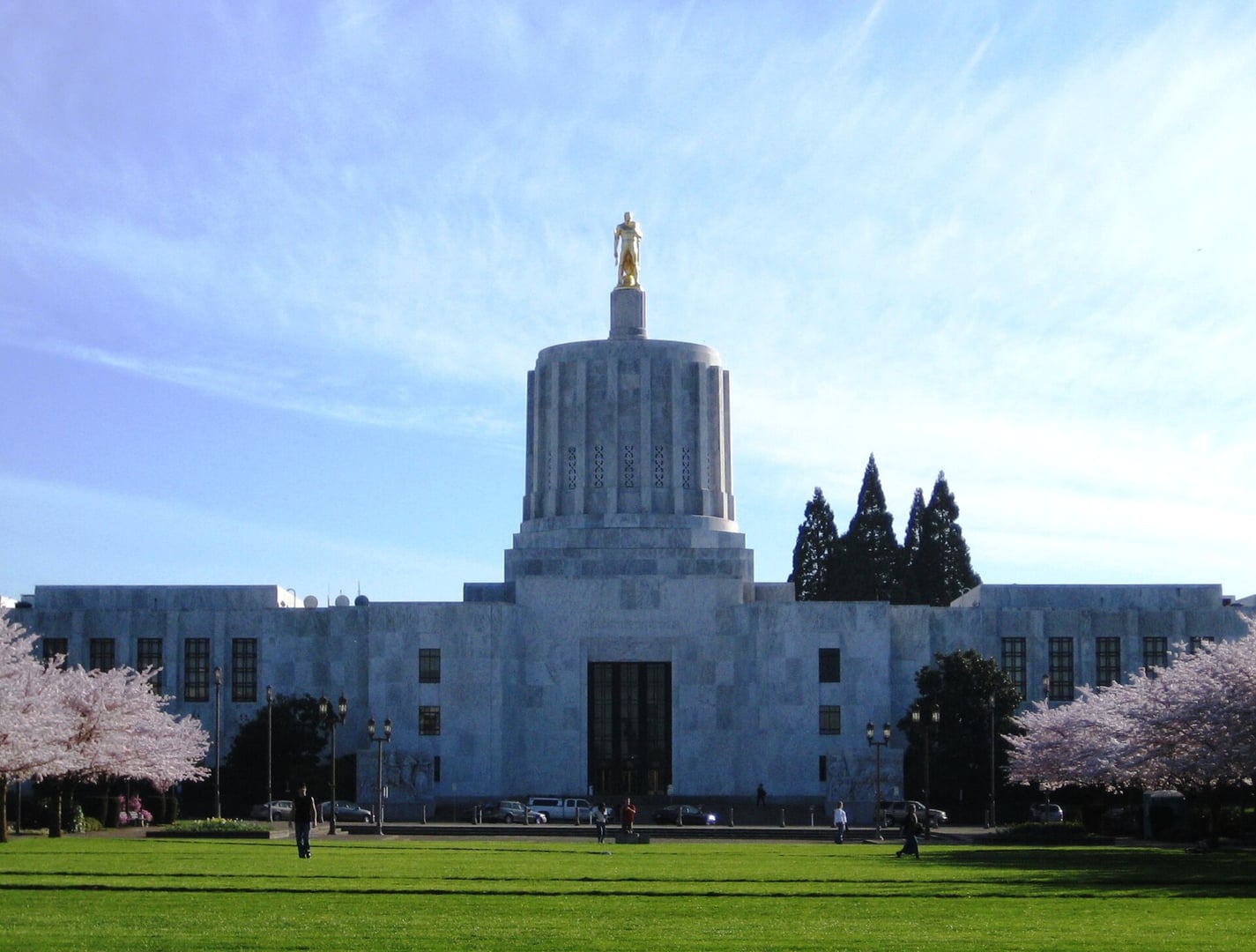 Front exterior of the 1938 Art Deco Oregon State Capitol in Salem, with cherry trees in bloom along the Capitol Mall and the gilt-bronze Oregon Pioneer statue atop the cupola.