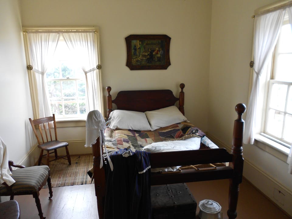 Restored keeper's-quarters bedroom on public display inside Yaquina Bay Lighthouse, Newport, Oregon: a wooden bedstead with a patchwork quilt, a bonnet and dress laid over the footboard, two chairs and curtained windows (photographed 17 September 201