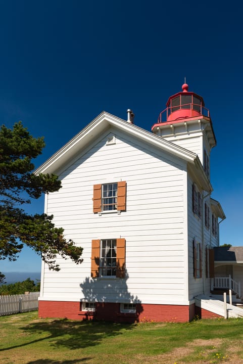 Gable-end (side) view of Yaquina Bay Lighthouse, Newport, Oregon: the white clapboard keeper's house with shuttered windows and a red brick foundation, and the red-roofed lantern tower rising behind the roof (photographed 9 September 2013). Photo: Ga