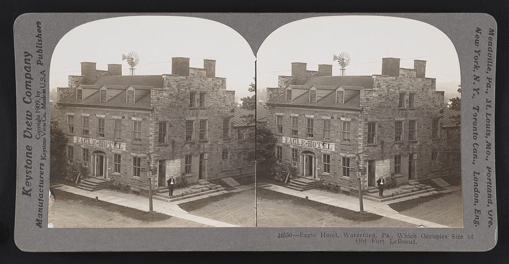A 1909 Keystone View Company stereograph of the Eagle Hotel in Waterford, Pennsylvania, captioned 'which occupies site of old Fort LeBoeuf,' showing the fieldstone building with its painted sign, a windmill, and a man standing on the front steps.