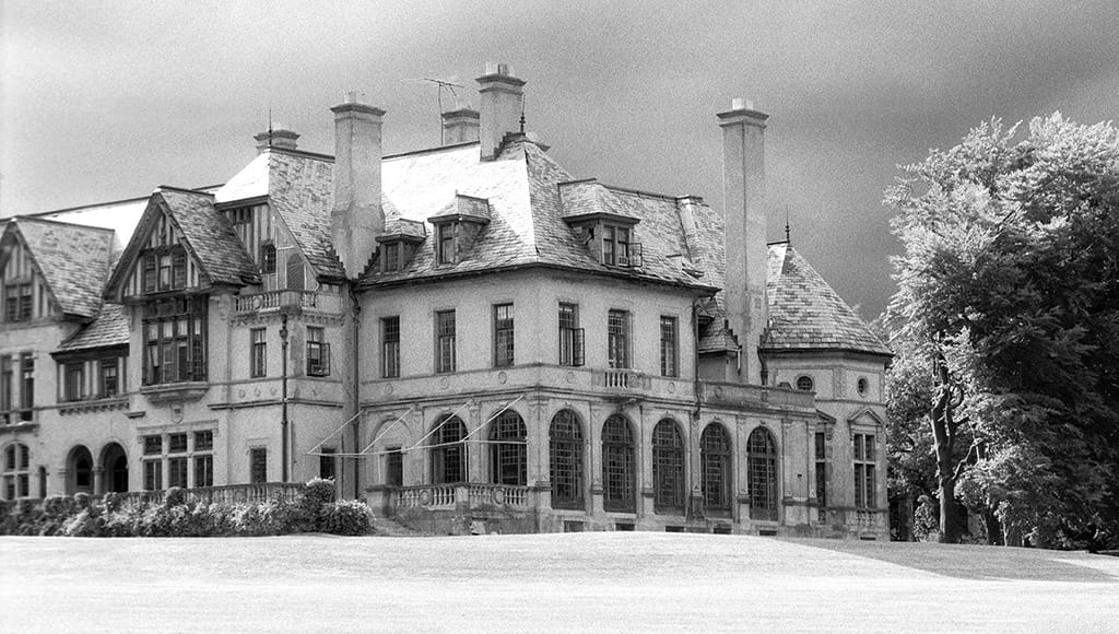 The arcaded garden front of Carey Mansion (Seaview Terrace) in Newport, Rhode Island, seen from the lawn, with arched ground-floor windows, tall chimneys, a slate roof and a half-timbered gable, in a black-and-white photograph from 1998.