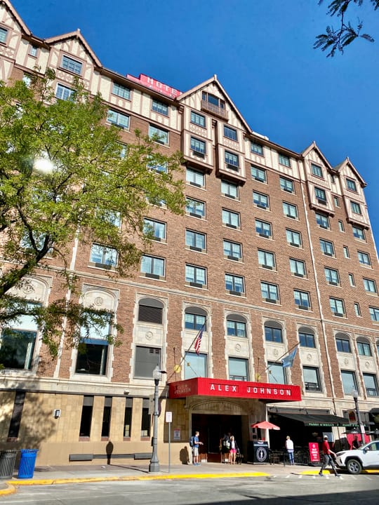 Street-level view of the hotel's main entrance and red 'ALEX JOHNSON' canopy, at the corner of St. Joseph Street and 6th Street, Rapid City, SD.