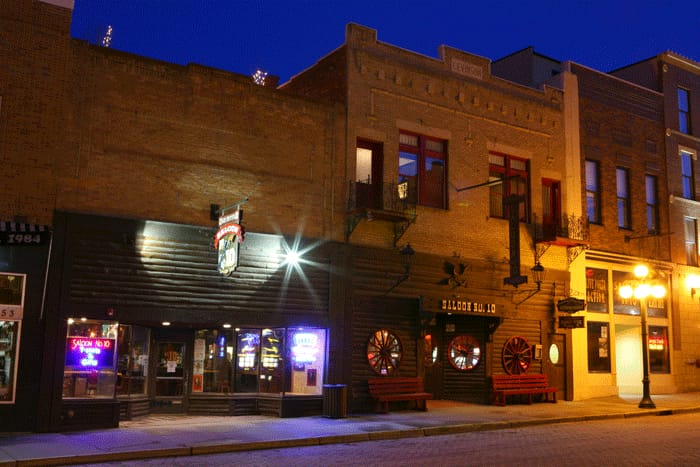 The log-clad front of the Old Style Saloon No. 10 at 657 Main Street in Deadwood, South Dakota, at dusk, with the 'Saloon No. 10' sign over the door, wagon-wheel panels and lit windows, in a photograph dated 2011.