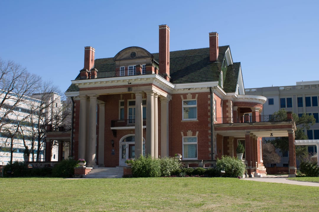 Thistle Hill (the Wharton-Scott House) in Fort Worth, Texas, seen at an angle across the lawn, with the two-storey columned portico, pedimented dormer, red-brick walls and curved side porch, in 2016.