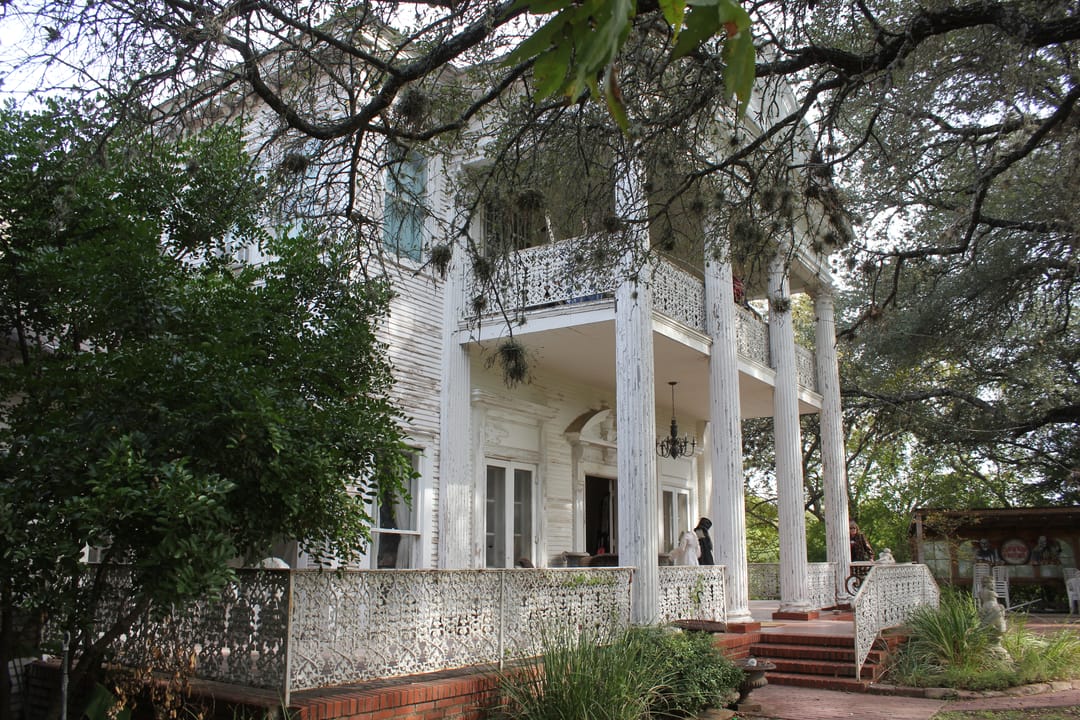 Daytime view of Victoria's Black Swan Inn's wraparound porch and columned facade, framed by live oaks.