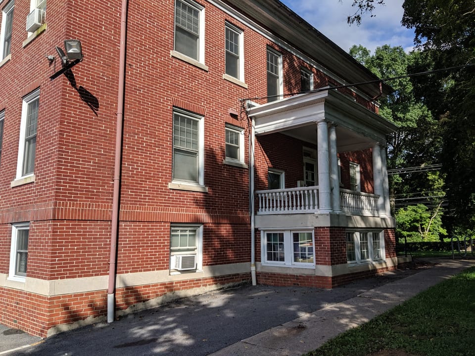 The side elevation of Miller Hall at Shepherd University, Shepherdstown, West Virginia, showing the red-brick wall and the end porch with its white balustrade, August 2019.