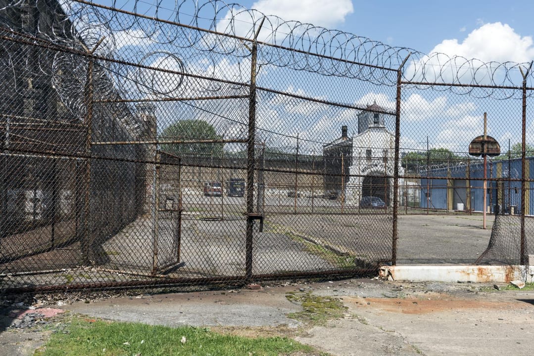An old exercise yard at the West Virginia State Penitentiary, enclosed by razor-wire fencing, with the cellblock building visible beyond.