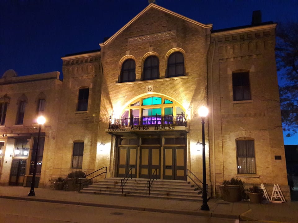 Front facade of The Grand Opera House lit at night before dawn, with 'GRAND OPERA HOUSE' carved above the entrance and colored light in the arched balcony window, October 2018.