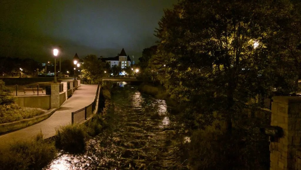 The Waukesha Fox River Riverwalk at night, with gaslit walkway path and downtown buildings illuminated in the distance, starting point of the Waukesha Ghost Walk