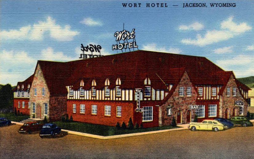 Colour-printed linen postcard (Curt Teich, 1942) of the Wort Hotel in Jackson, Wyoming, as first built: a Tudor-style stone, brick and half-timber building with two rooftop 'Wort Hotel' signs, BAR and CAFE signs and period cars, titled 'Wort Hotel - 
